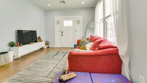 A modern living room with a red sofa, a flat-screen TV, and a large window with sheer curtains. - Work & Play: Colorful Home in Heart of NOLA! (New Orleans)