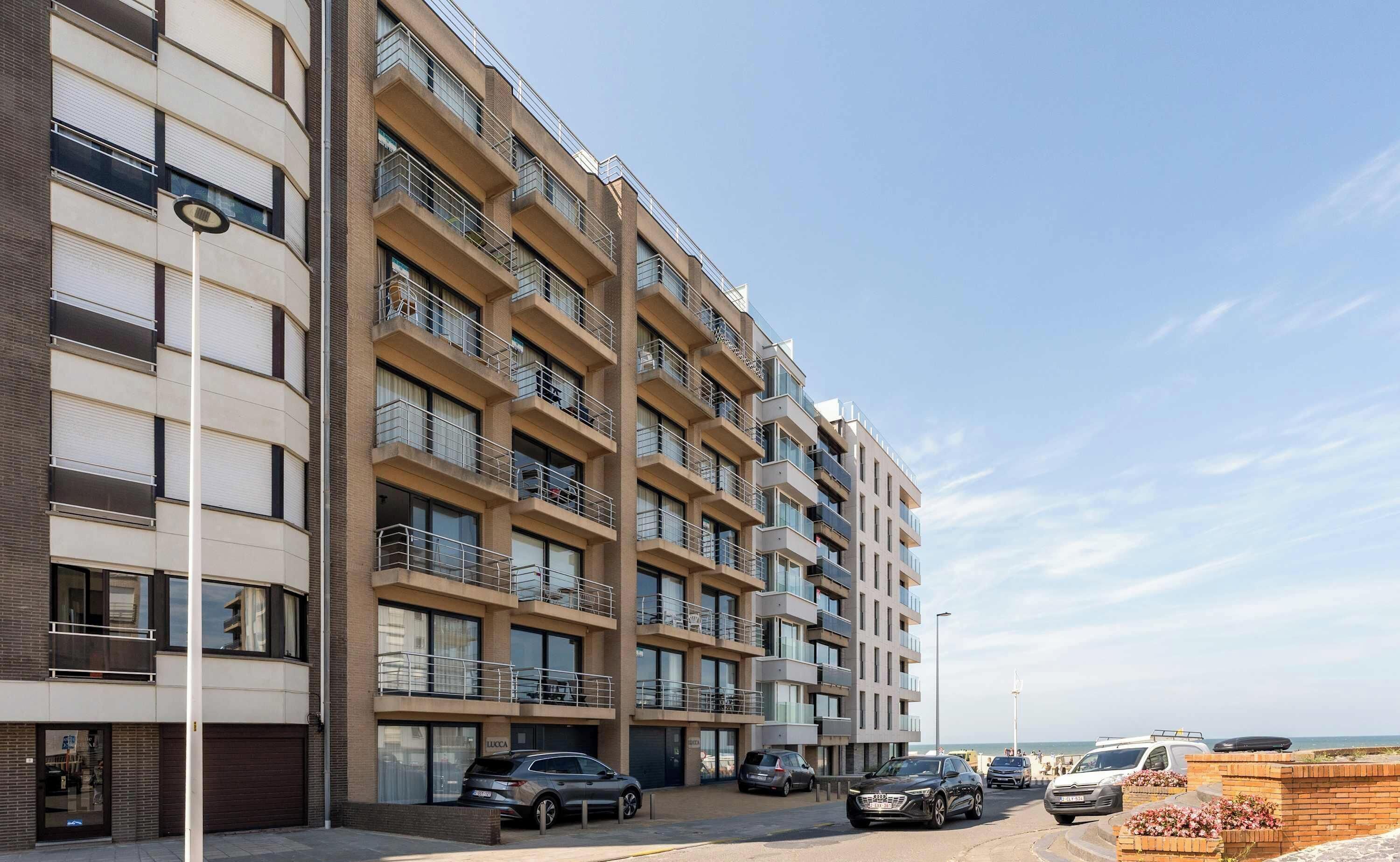 A multi-story residential building with balconies, a parking area, and a view of the sea.