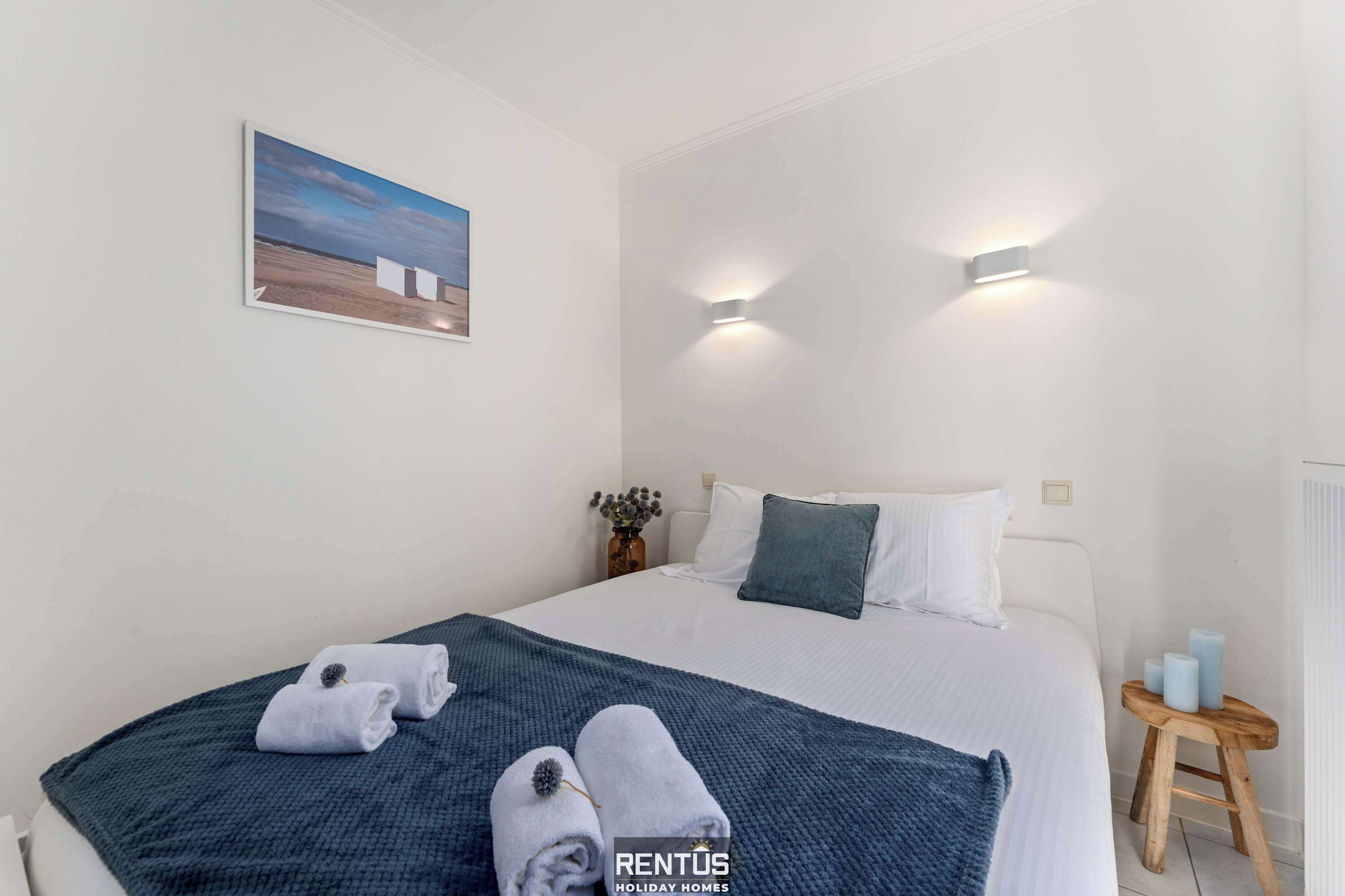 Ein Schlafzimmer mit einem Bett, zwei Wandleuchten, einem Gemälde, einem kleinen Tisch mit Kerzen und einer blauen Decke.