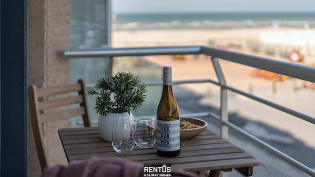 A balcony with a wooden table, a bottle of wine, two glasses, a bowl of snacks, and a potted plant.