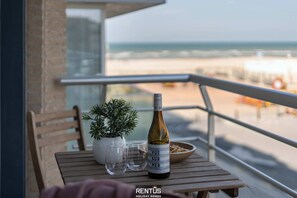 Un balcón con una mesa de madera, una botella de vino, dos copas, un cuenco de aperitivos y una planta en maceta.