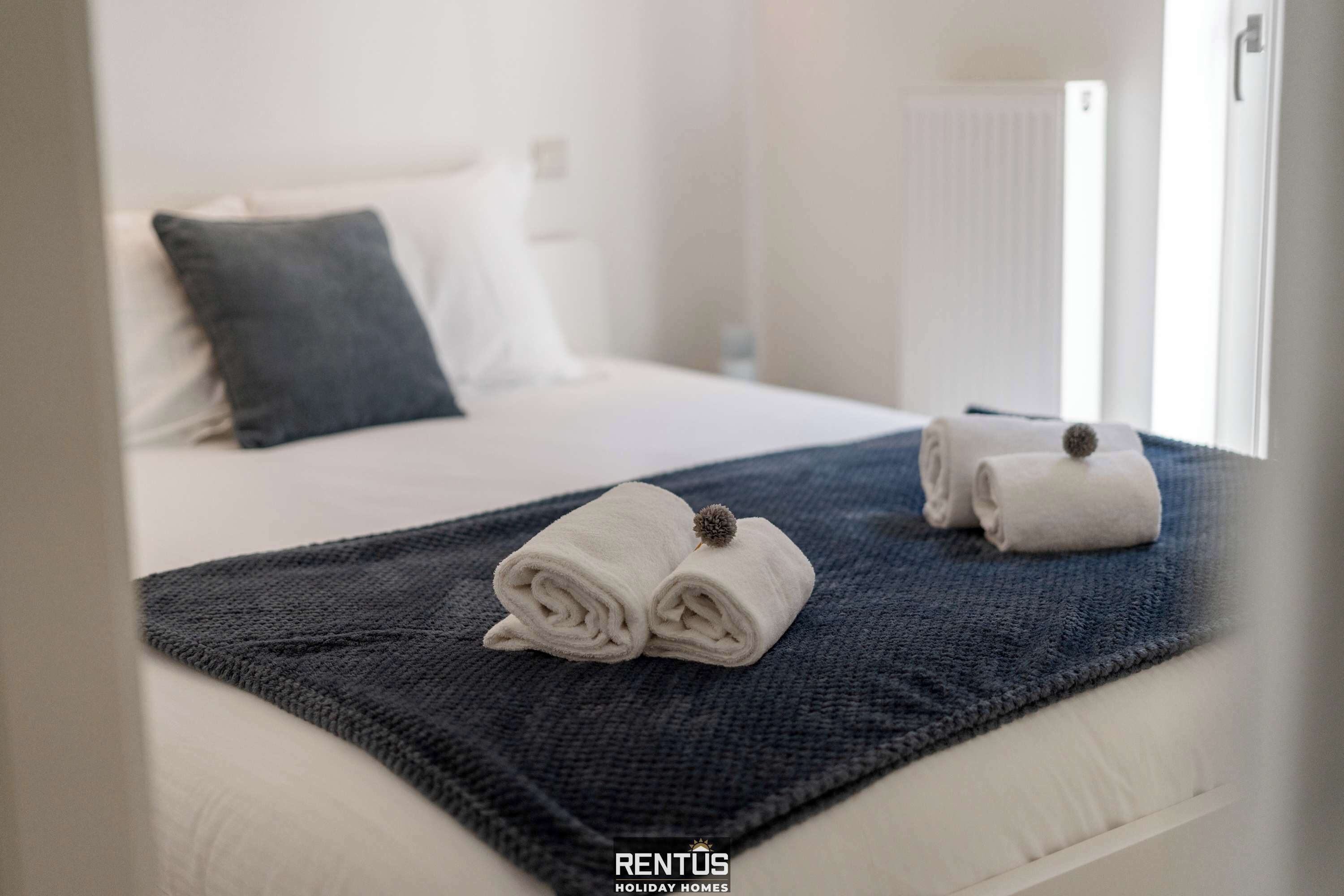 Ein ordentlich bezogenes Bett mit zwei aufgerollten Handtüchern und einer dunkelblauen Decke, einem weißen Kopfkissen und einem weißen Kopfteil.