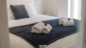 A neatly made bed with two rolled towels and a dark blue blanket, a white pillow, and a white headboard.