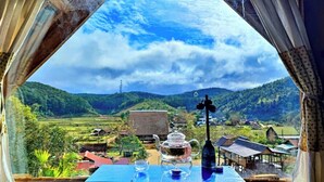 A table with a teapot, cup, and saucer, a bottle, and a view of a rural landscape with houses and hills. - To Chim Ri Homestay Mang Đen (Mang Den)