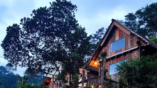 A rustic wooden building with multiple levels, surrounded by lush greenery and trees. - To Chim Ri Homestay Mang Đen (Mang Den)