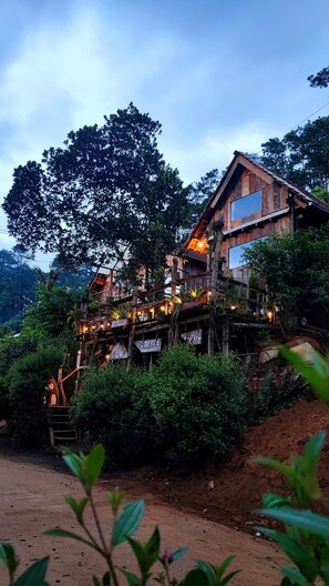A rustic wooden building with multiple levels, surrounded by lush greenery and trees. - To Chim Ri Homestay Mang Đen (Mang Den)