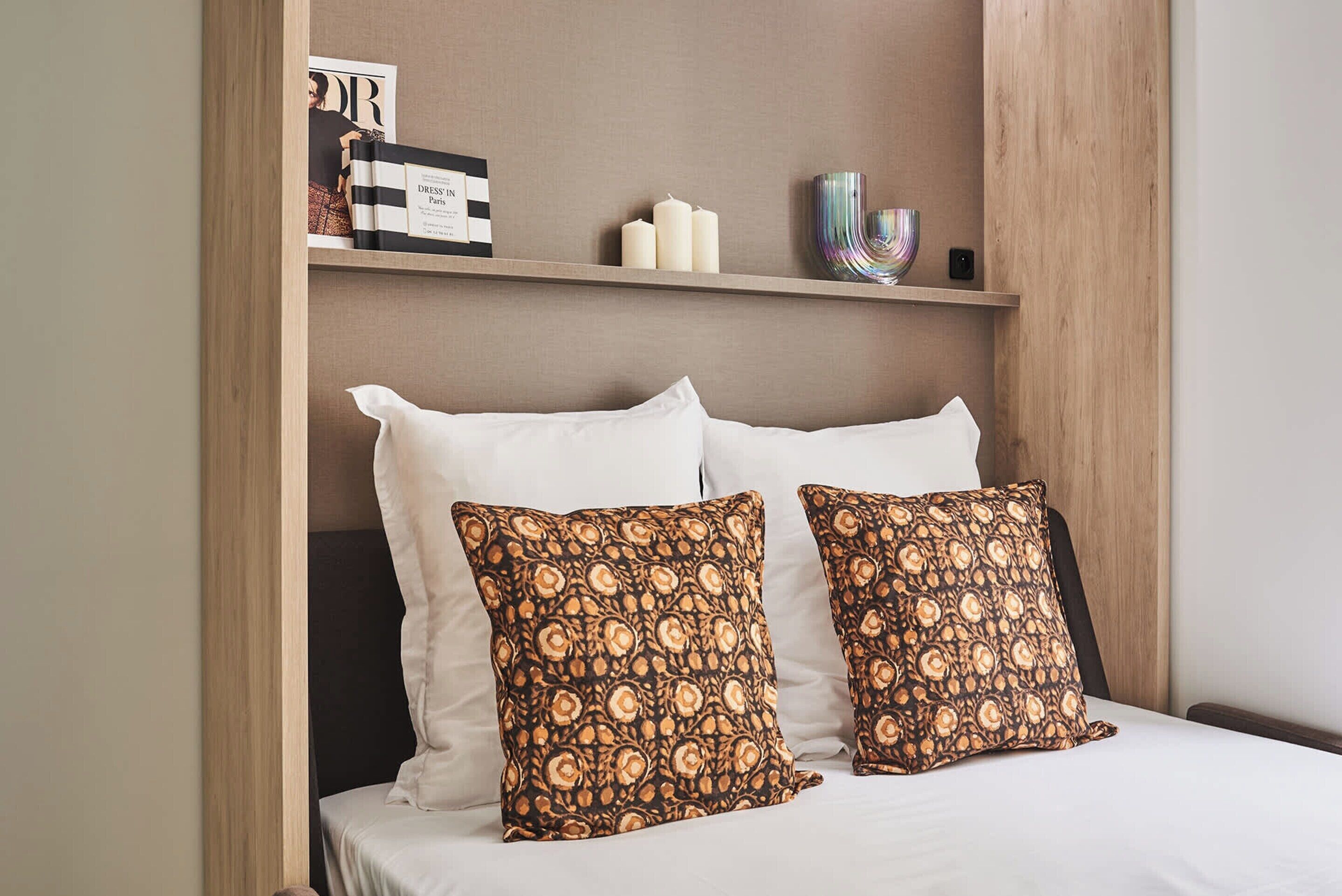 Un lit bien fait, avec du linge de lit blanc et deux coussins à motifs, une étagère avec un magazine, des bougies et un bol décoratif.