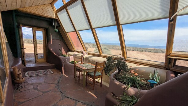 A cozy, sloped-roofed room with a stone floor, a dining area with chairs and a table, and a view of a desert landscape through large windows. - SolShip, An Original Earthship in Tres Piedras (Tres Piedras)