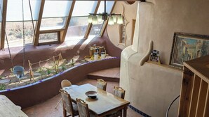 A cozy, uniquely designed room with a sloped ceiling, a dining table with chairs, a cushioned bench, and a small garden area visible through a window. - SolShip, An Original Earthship in Tres Piedras (Tres Piedras)