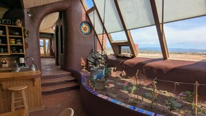 A rustic kitchen with a wooden bar and a sink, a dining area with a bench and chairs, and a garden area with plants and a swing. - SolShip, An Original Earthship in Tres Piedras (Tres Piedras)