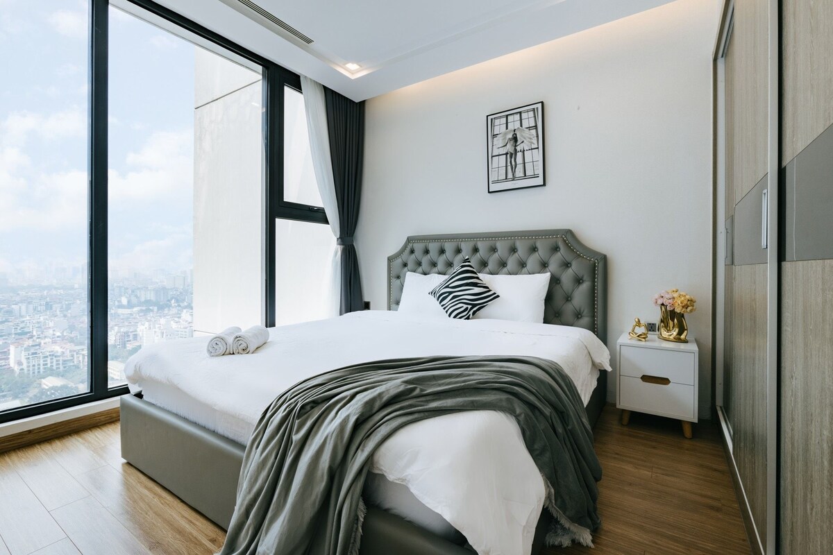 A modern bedroom with a large bed, a nightstand, a framed artwork, and a view of the city through a large window.