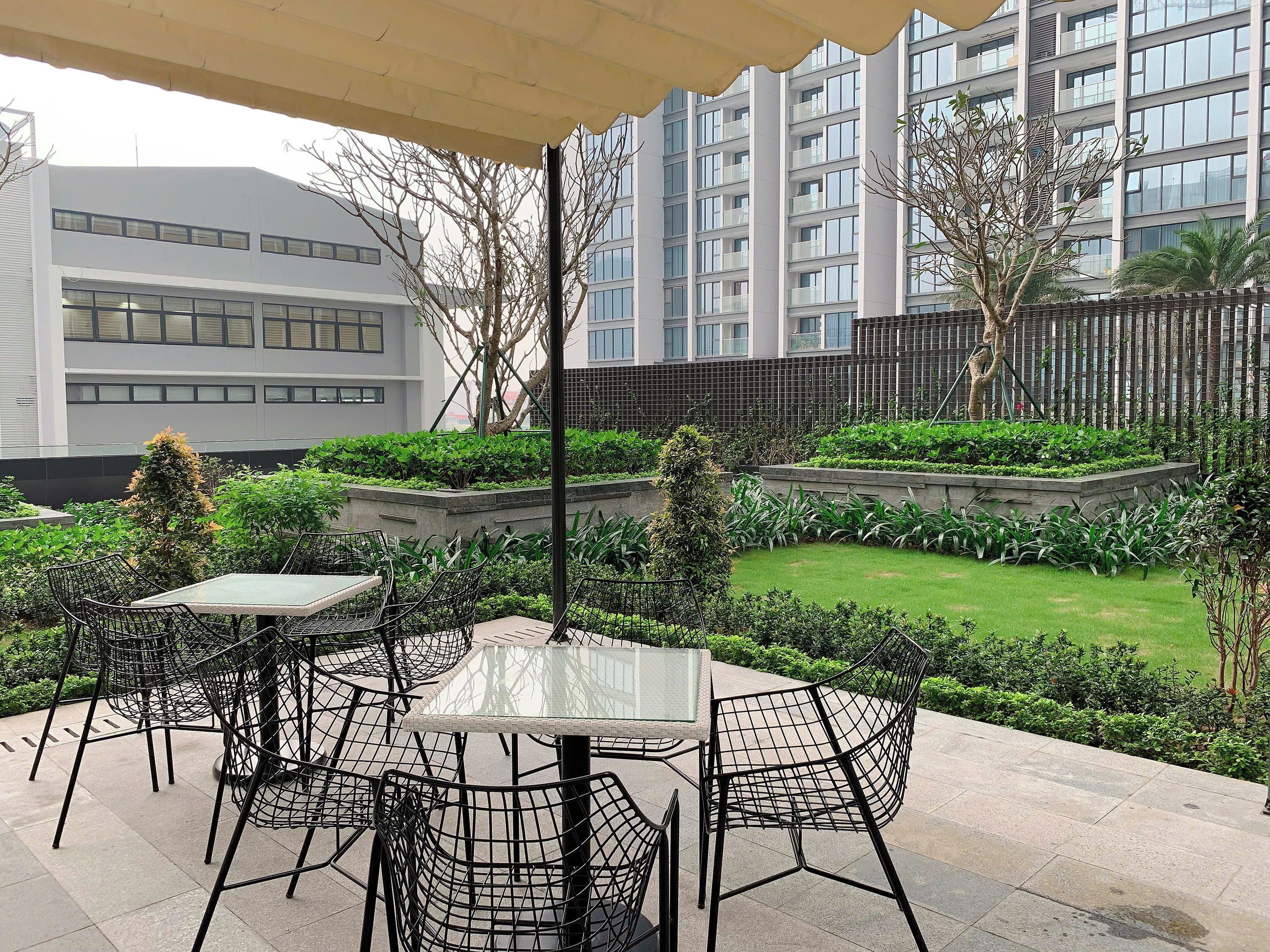 An outdoor patio area with tables and chairs, surrounded by greenery and a view of modern buildings.