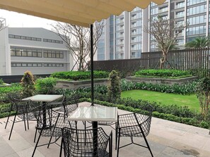 Een terras buiten met tafels en stoelen, omringd door groen en een uitzicht op moderne gebouwen.