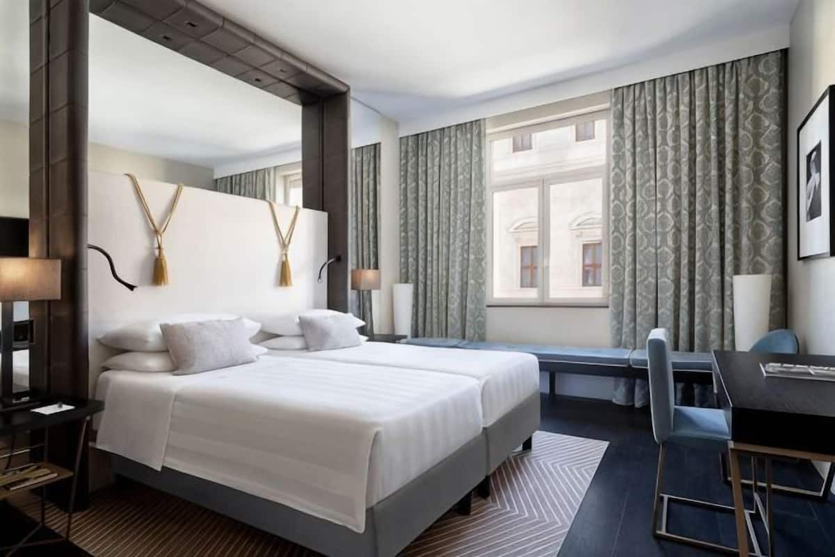 Hotellihuoneessa on suuri sänky, työpöytä ja tuoli. Ikkunoissa on verhot ja työpöydällä lamppu.