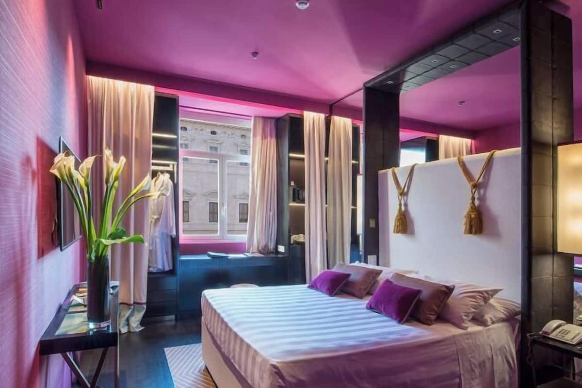 Moderni hotellihuone, jossa on suuri sänky, yöpöytä, peili ja ikkunasta avautuva näkymä kaupungin ylle.