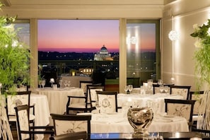 Un restaurante con mesas dispuestas para comer, amplias ventanas con vista a un edificio e iluminación decorativa.