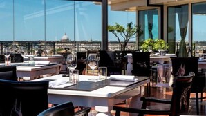 A rooftop restaurant with tables set for dining, large windows offering a view of a dome structure, and potted plants on the tables. - Sina Bernini Bristol, Autograph Collection (Rome)