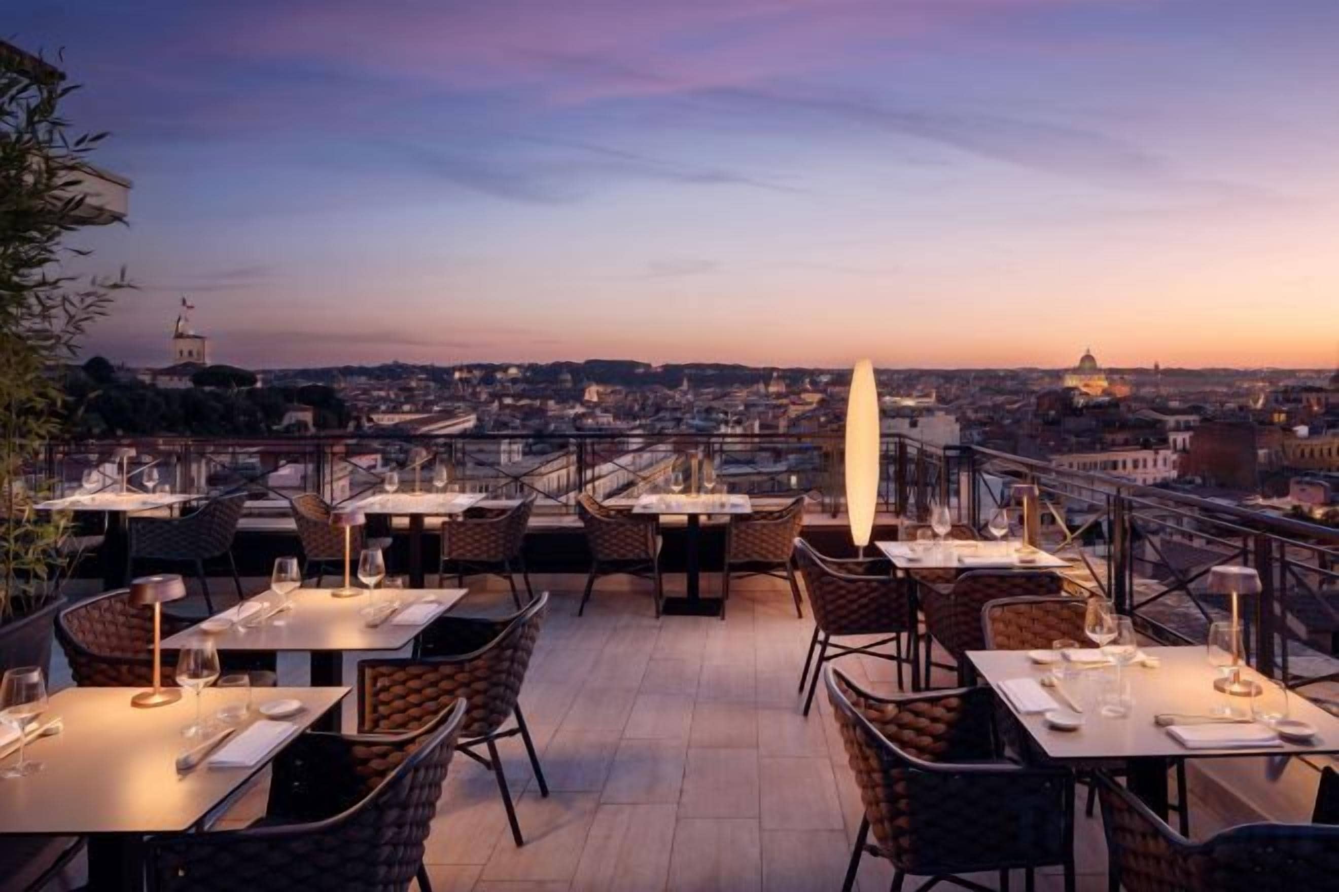 An outdoor dining area with tables and chairs, overlooking a cityscape at sunset.