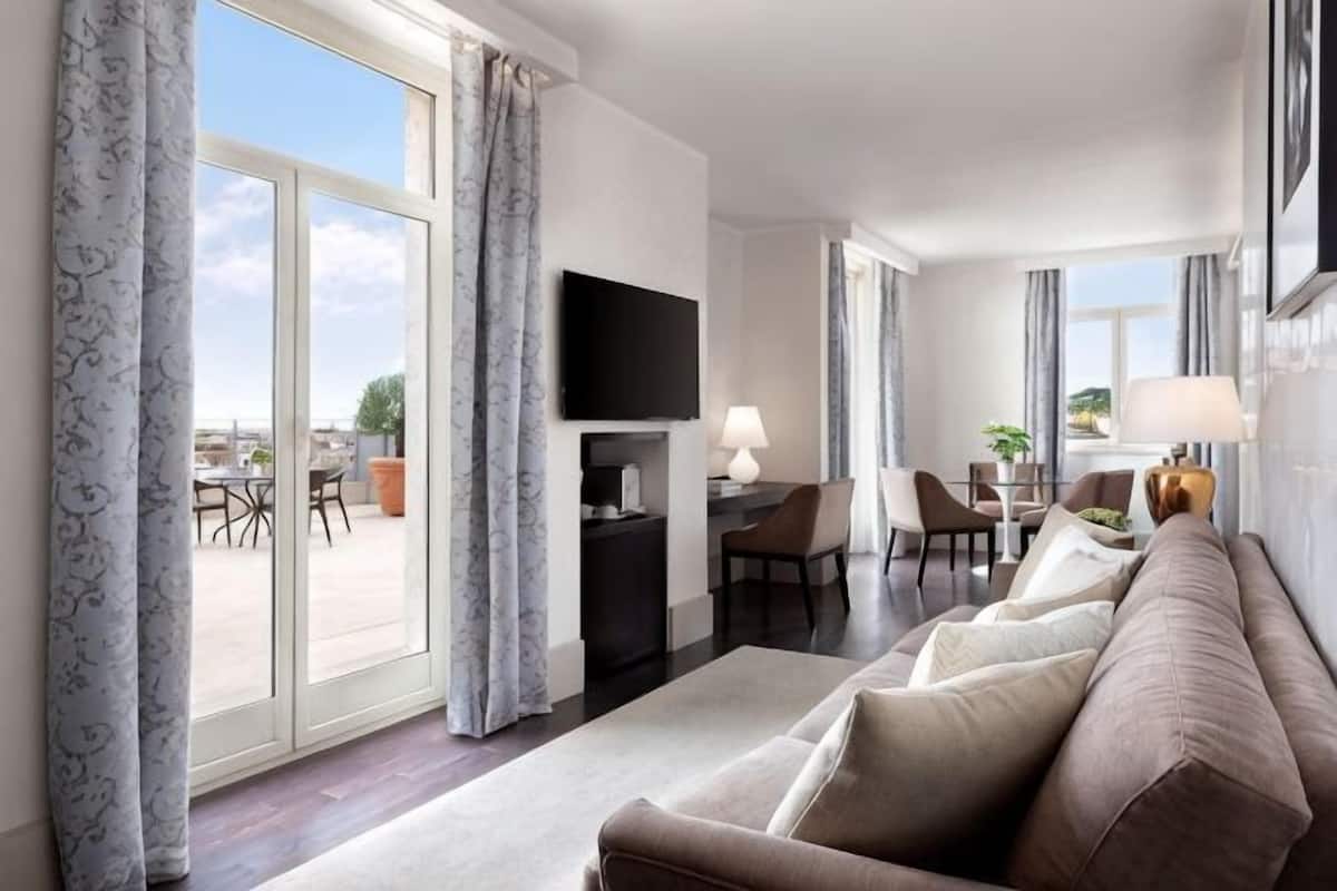 Hotellihuone, jossa on sohva, televisio, ruokailutila ja suuret ikkunat.