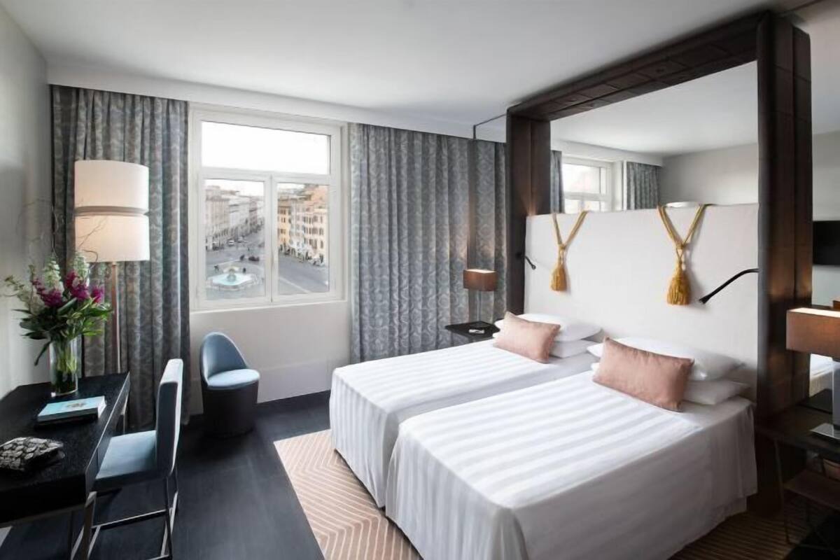Hotellihuone, jossa on kaksi sänkyä, työpöytä tuolilla, lamppu ja suuret ikkunat, joista on näkymä kaupunkiin.