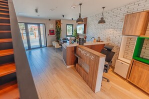 Ein Empfangsbereich mit Holzausstattung und dem Schriftzug 'BOCOBAY'. Zwei Stühle und ein kleiner Tisch mit einer Pflanze stehen auf dem Schreibtisch. Der Raum ist modern gestaltet, mit einer Ziegelwand und einem großen Fenster.