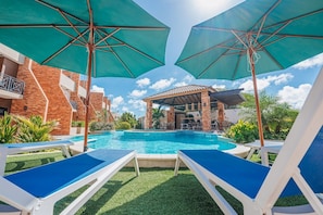 A swimming pool with lounge chairs and umbrellas, surrounded by brick buildings and greenery. - LUX 3BR House Pool Gym 7-Min Walk Palm Beach - Porta Al Sole 6 by Bocobay (Noord)