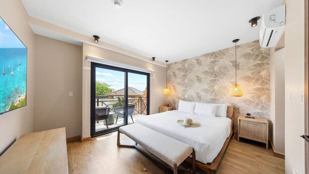 A bedroom with a large bed, a bench, a nightstand, a wall-mounted air conditioner, and a view of the ocean through a sliding door. - LUX 3BR House Pool Gym 7-Min Walk Palm Beach - Porta Al Sole 6 by Bocobay (Noord)