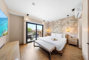 Un dormitorio con una cama grande, un banco, una mesita de noche, un aire acondicionado instalado en la pared y vistas al océano a través de un ventanal corredero.