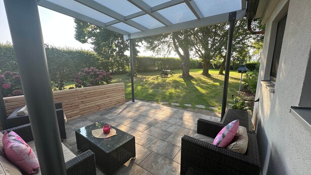 Un patio couvert aménagé avec du mobilier d’extérieur, un banc en bois et une vue sur un jardin bien entretenu.