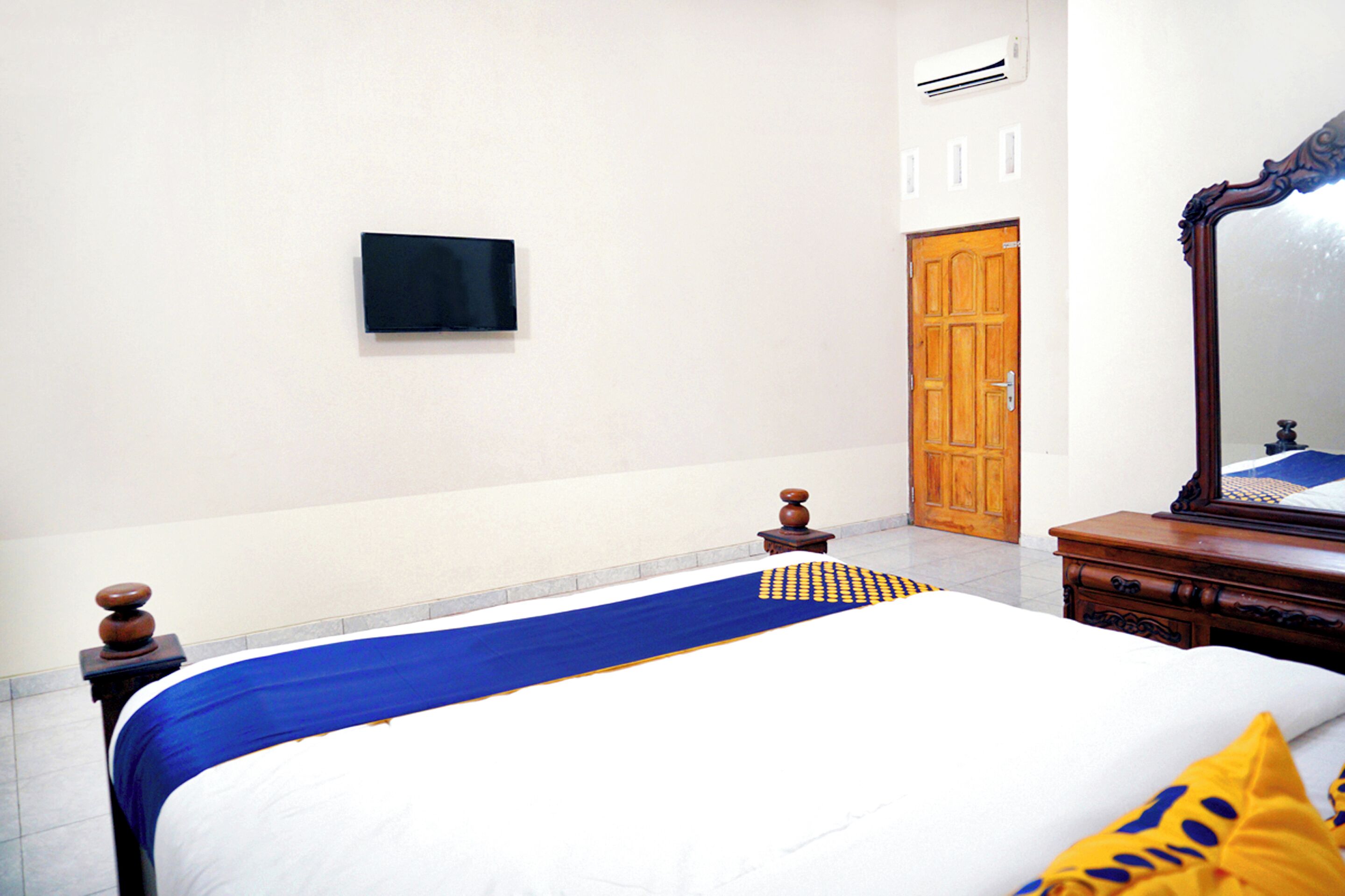 A bedroom with a bed, a wooden dresser, a mirror, a wardrobe, and a wall-mounted TV.