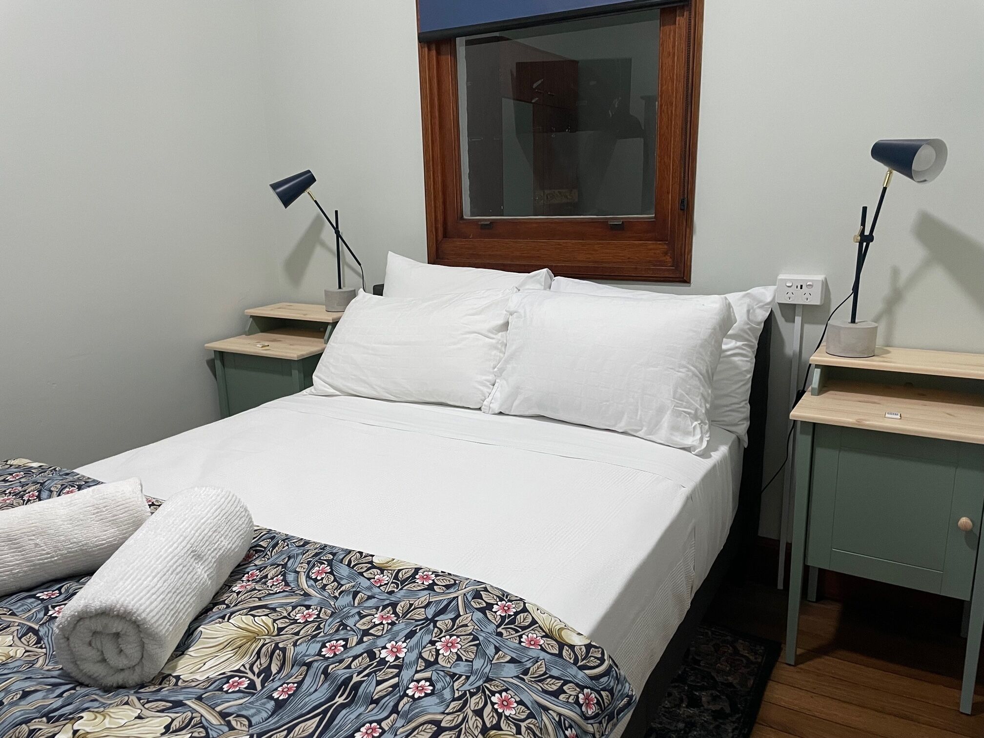 A neatly made bed with white linens and a floral-patterned duvet, two bedside tables with lamps, a window with a wooden frame, and a power outlet on the wall.