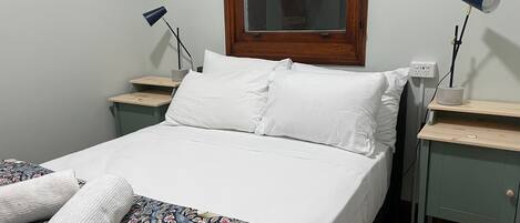 A neatly made bed with white linens and a floral-patterned duvet, two bedside tables with lamps, a window with a wooden frame, and a power outlet on the wall.