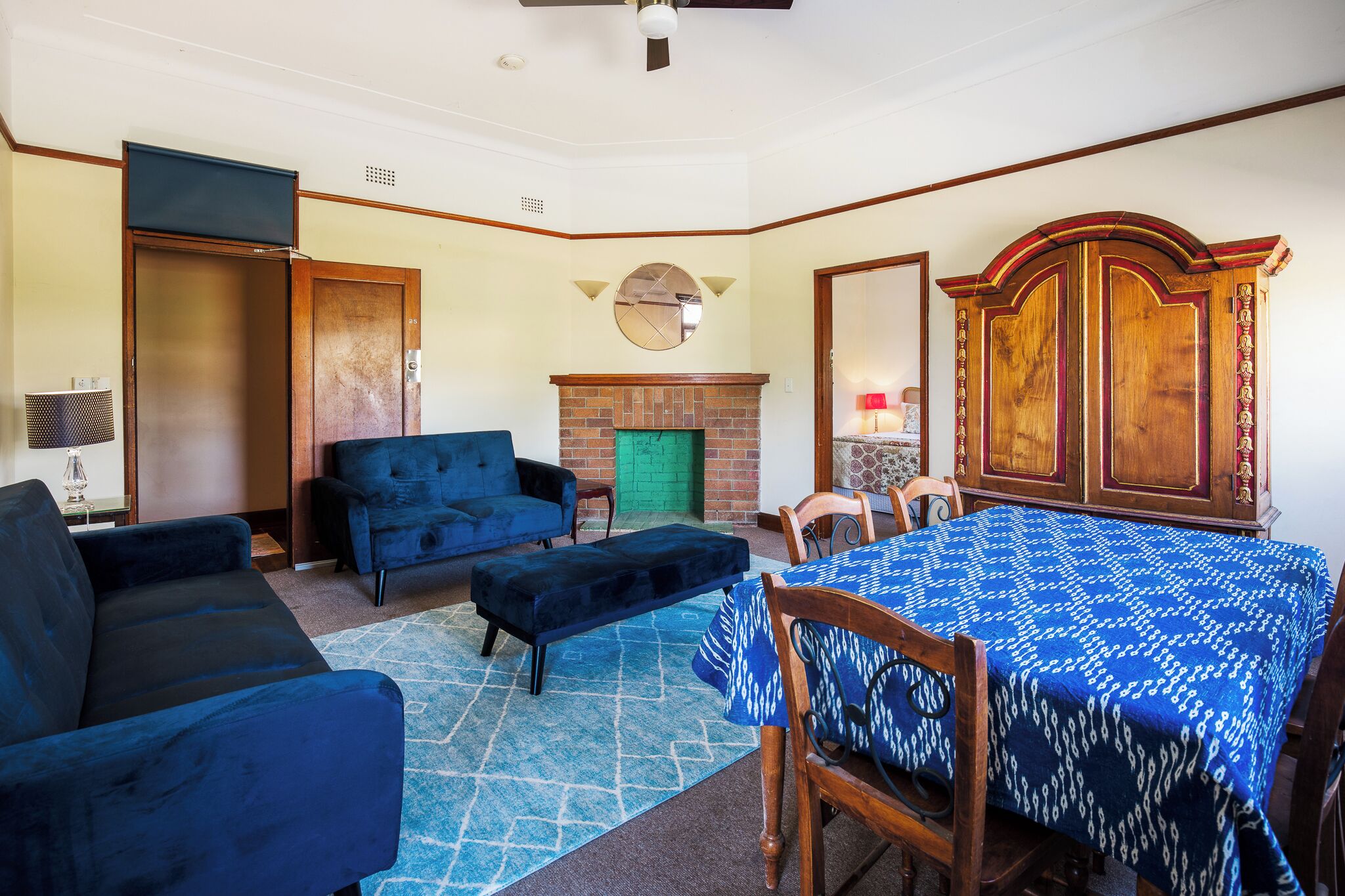 A room with a blue sofa, a matching ottoman, a dining table with a patterned tablecloth, a brick fireplace, and a large wooden cabinet with decorative panels.