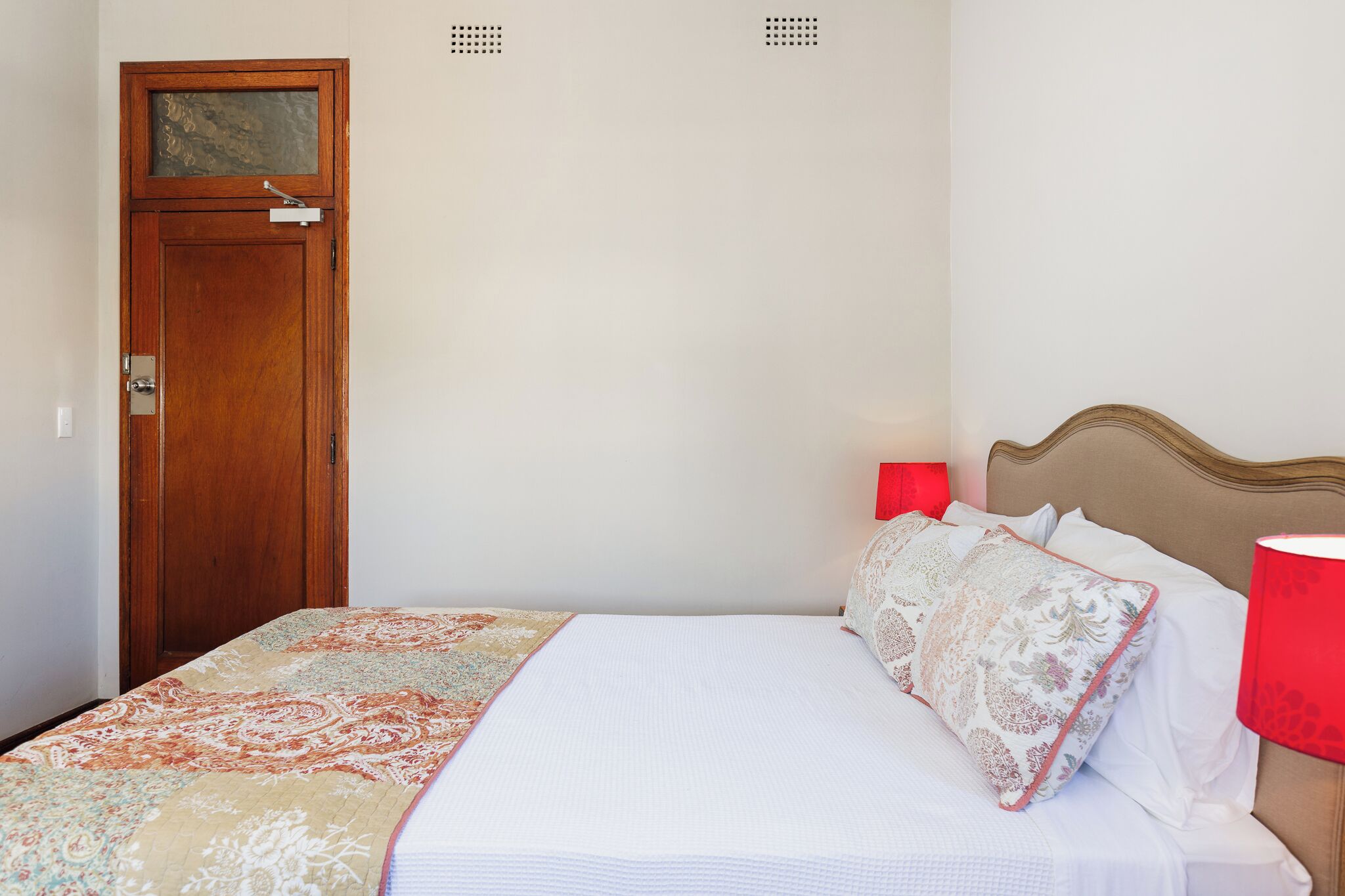 A bedroom with a wooden door, a bed with white bedding and patterned pillows, and a red lamp on the nightstand.