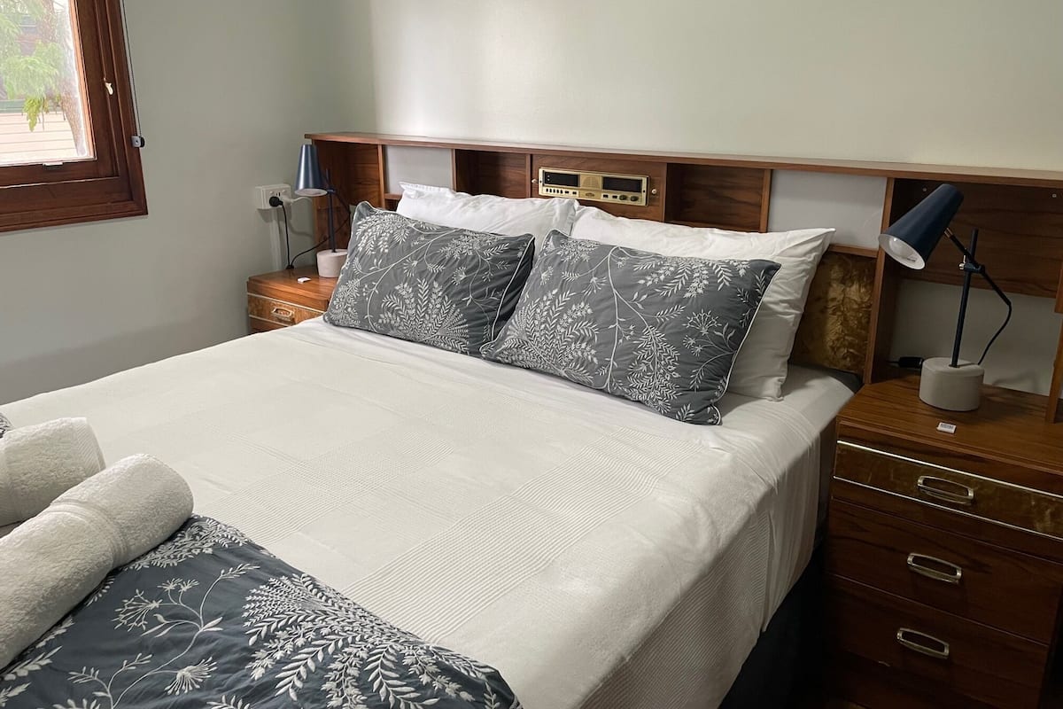 A neatly made bed with a white and gray floral bedspread, two pillows with similar patterns, a wooden nightstand with a lamp, and a radio on the wall.