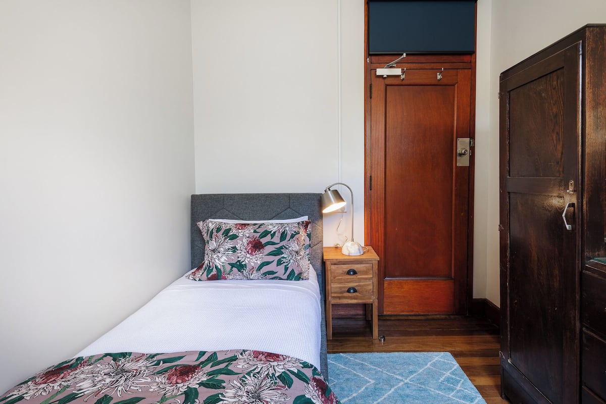 A single bed with a floral bedspread, a bedside lamp, a wooden nightstand, and a wardrobe.