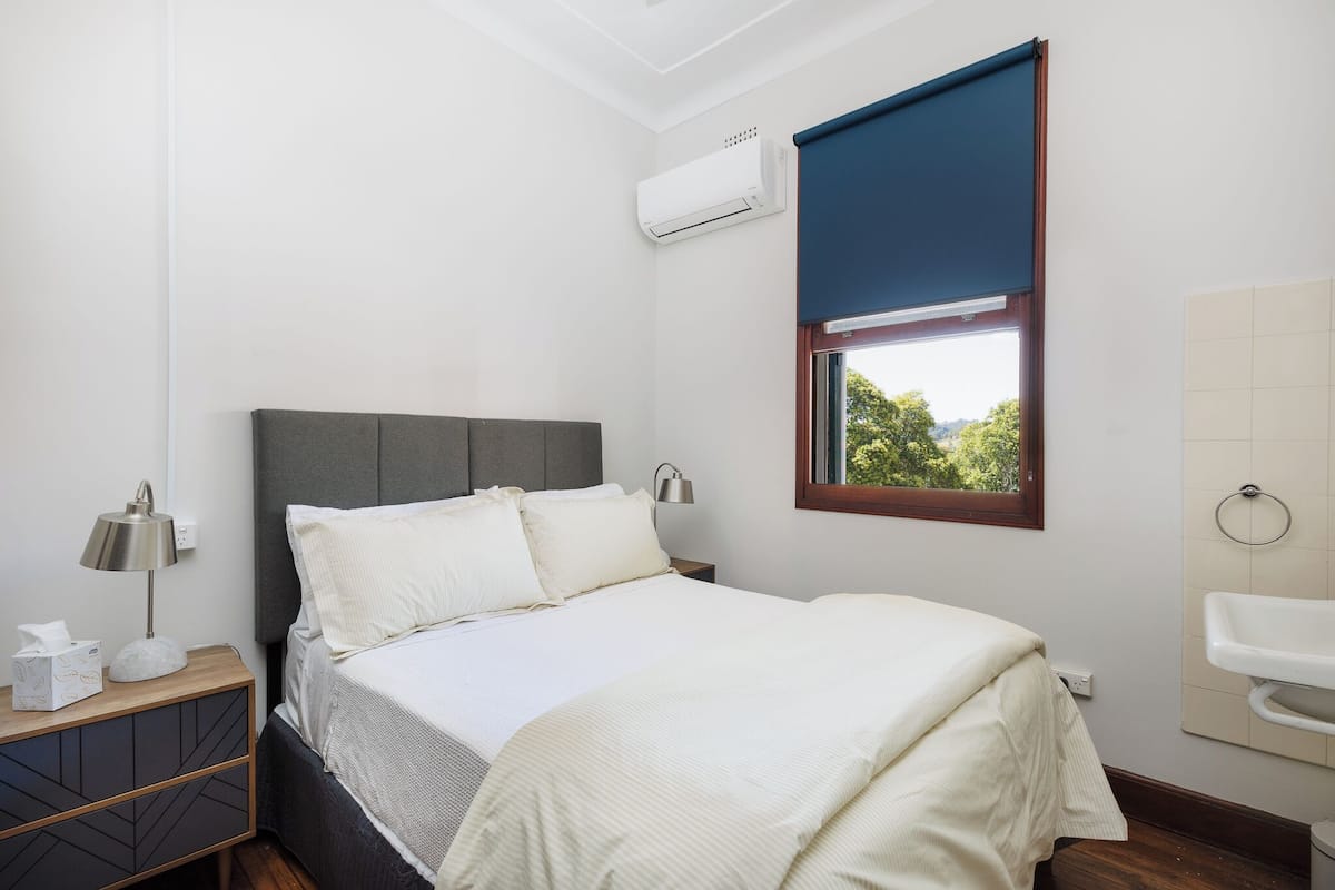 A bedroom with a bed, bedside lamp, air conditioning unit, window with view, and a nightstand with a lamp and tissues.