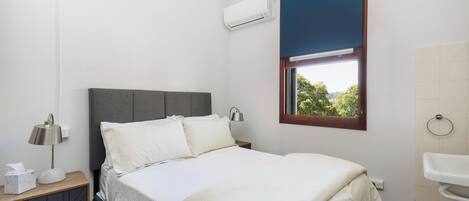 A bedroom with a bed, bedside lamp, air conditioning unit, window with view, and a nightstand with a lamp and tissues.