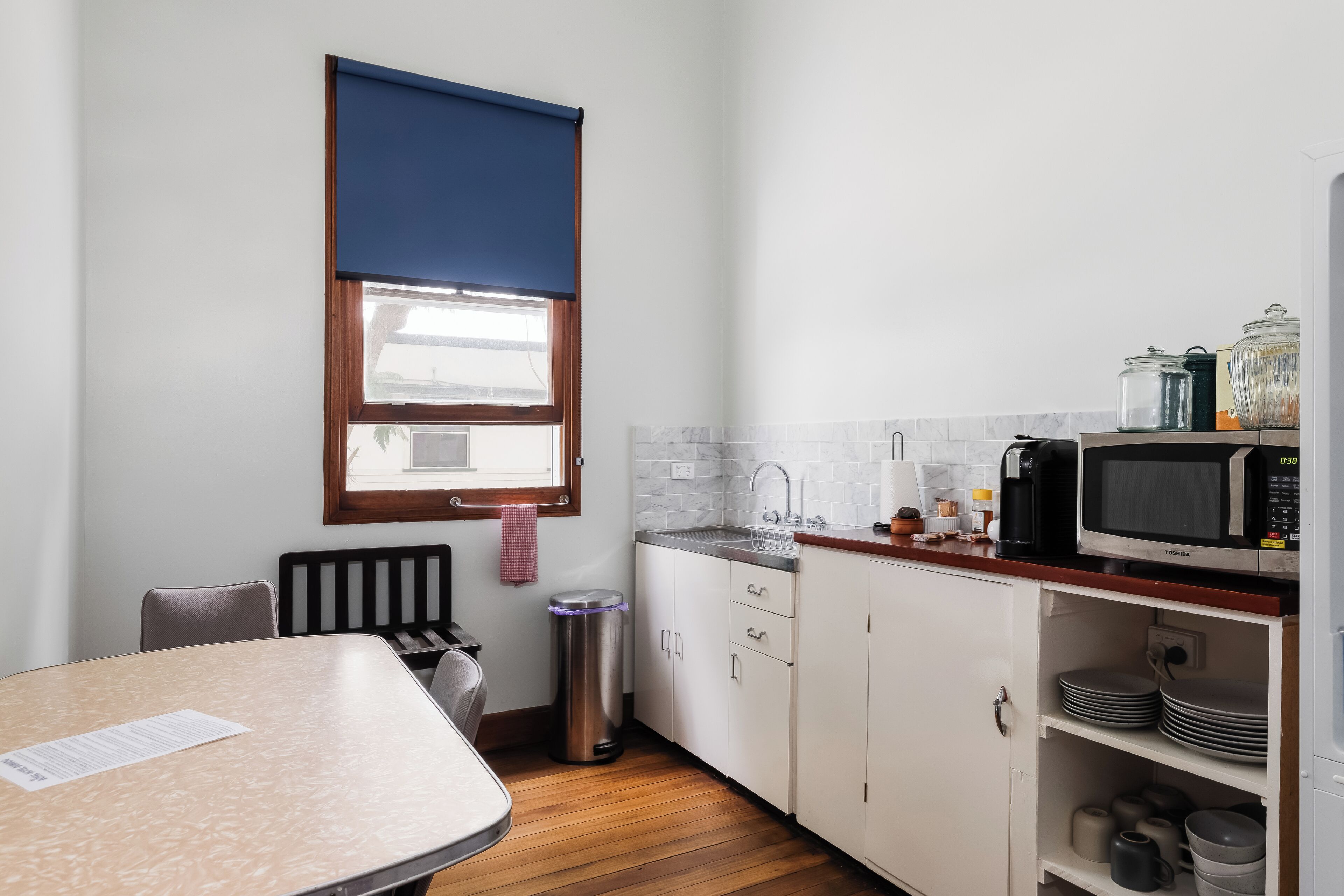 Una cocina compacta con área de comedor, una ventana con persiana azul, un bote de basura de acero inoxidable y un microondas en la encimera.