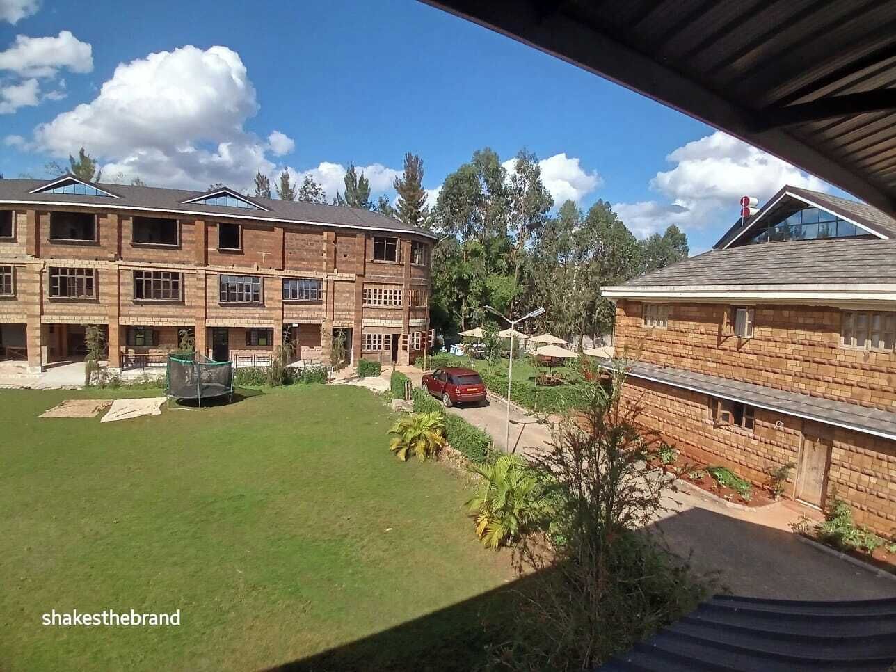 Una zona residencial con dos edificios de ladrillo, uno con balcón y otro con tejado plano. Un coche rojo estacionado en la calle, y un pequeño jardín con palmeras.