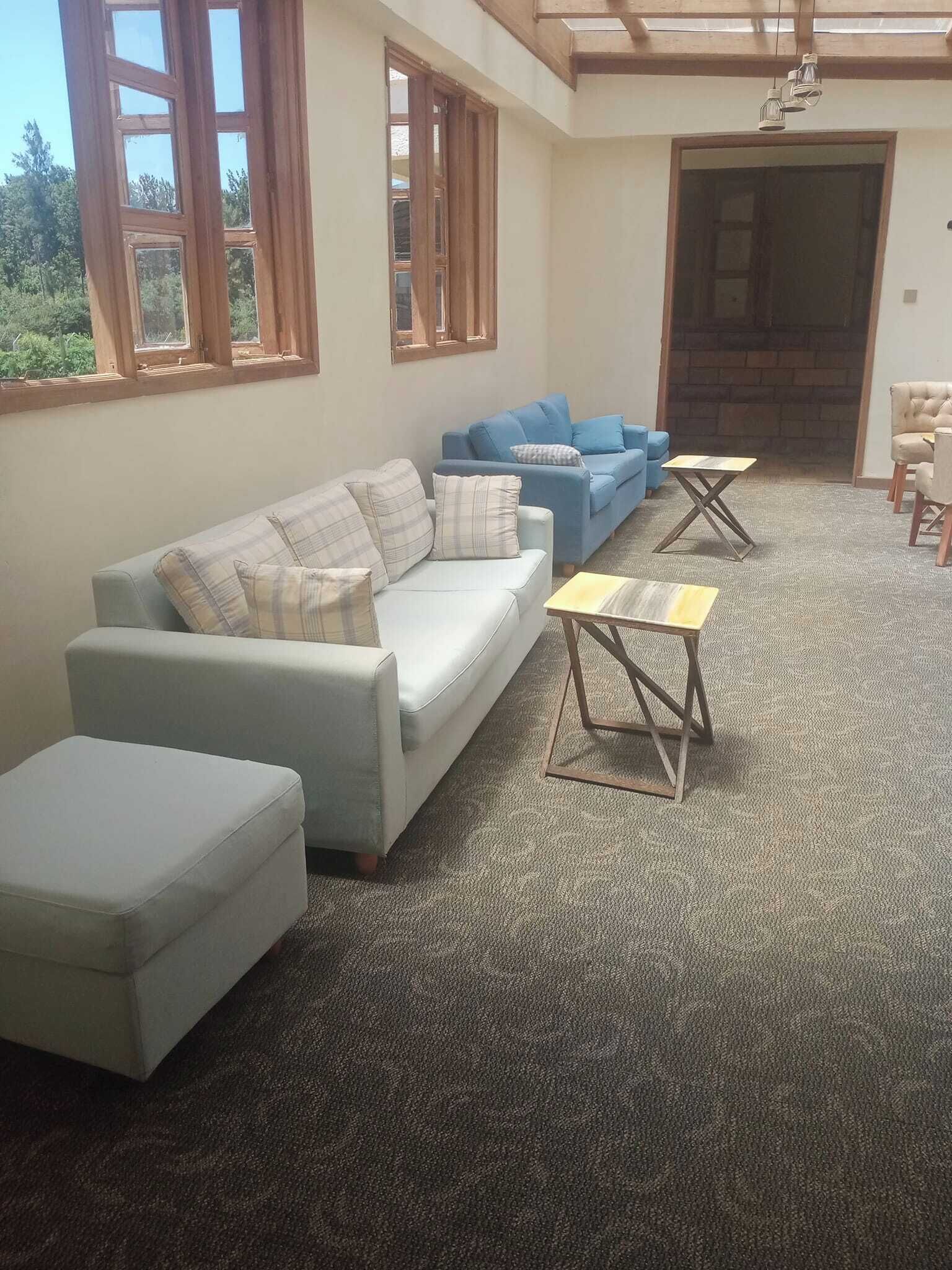 A living room with a light blue sofa, a blue sofa, and a beige armchair. There are two small tables and a large window with wooden frames.