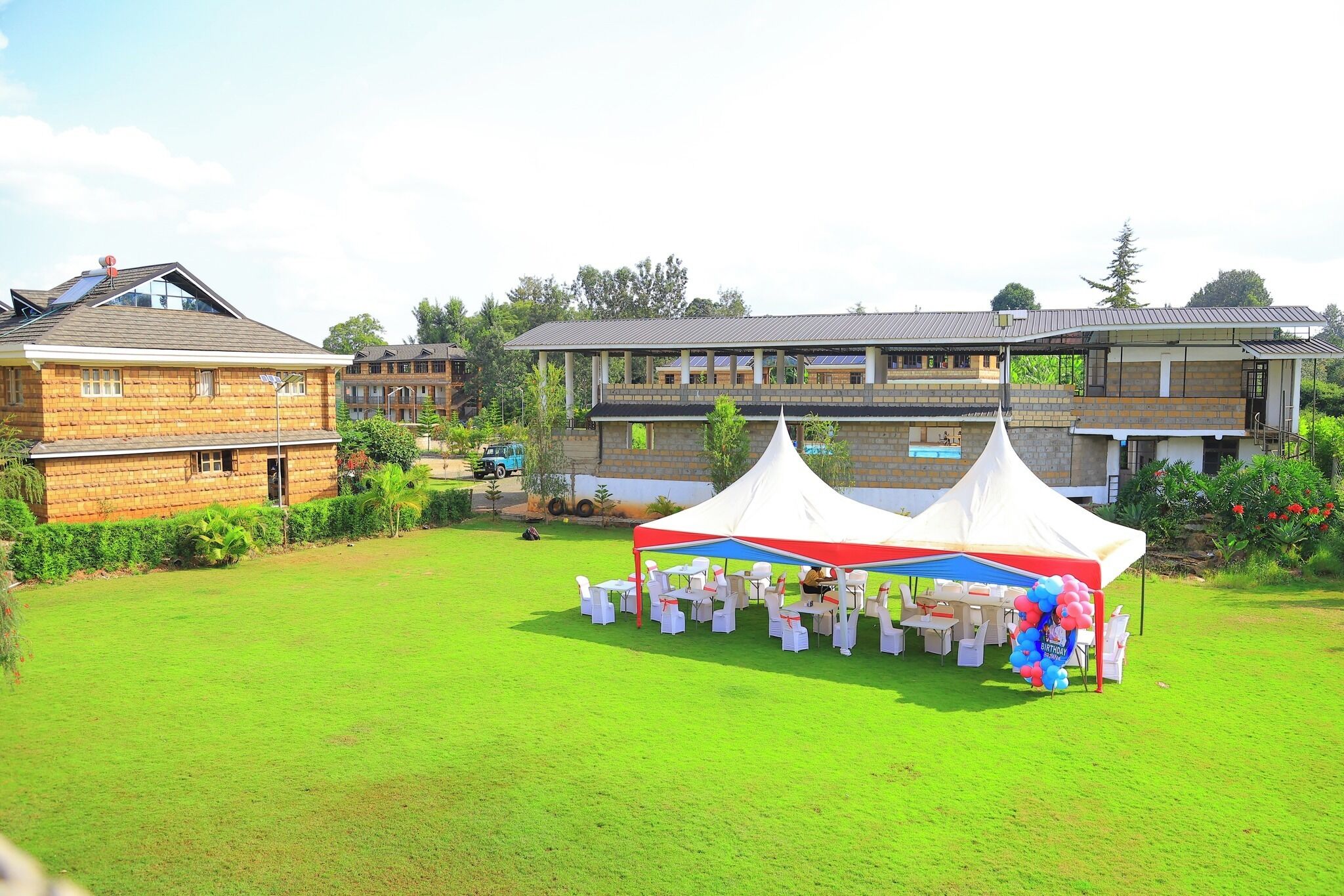 A residential area with a large house featuring a balcony, a white tent with a birthday banner, and a well-maintained lawn.