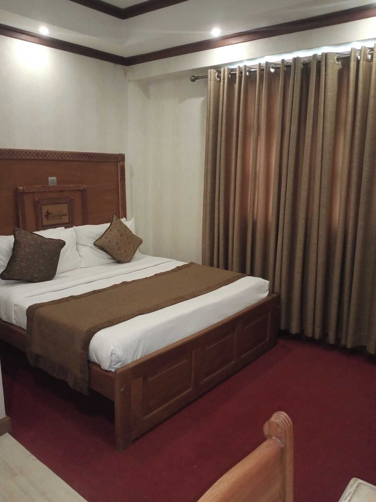 Una habitación de hotel con una cama de madera, ropa de cama blanca, un plaid marrón y una alfombra roja.