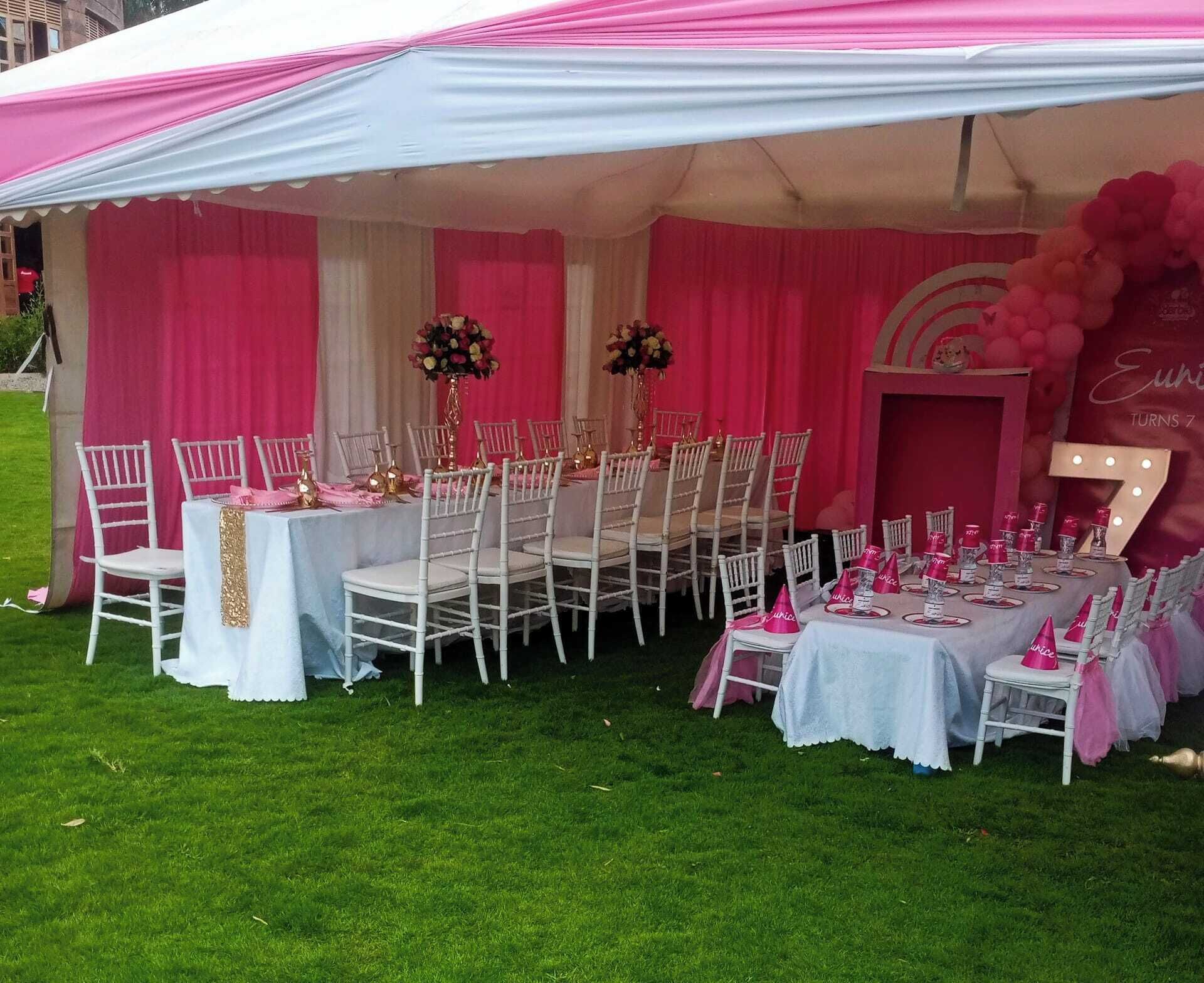 A tented outdoor event space with tables set for a celebration, featuring white chairs and pink decorations.