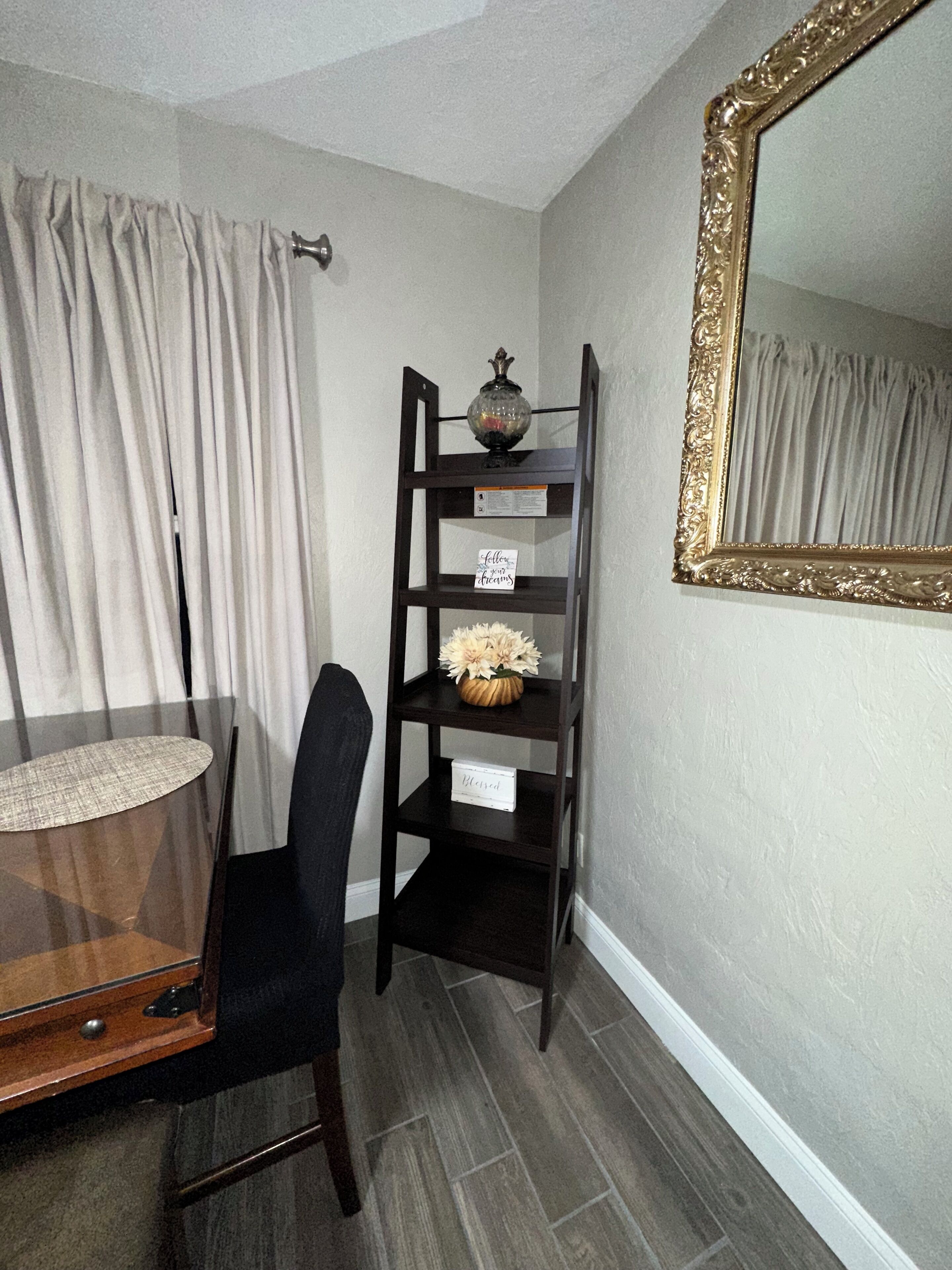 A room with a dark wooden chair, a small table with a lamp, a decorative shelf with a vase, and a mirror with a gold frame.