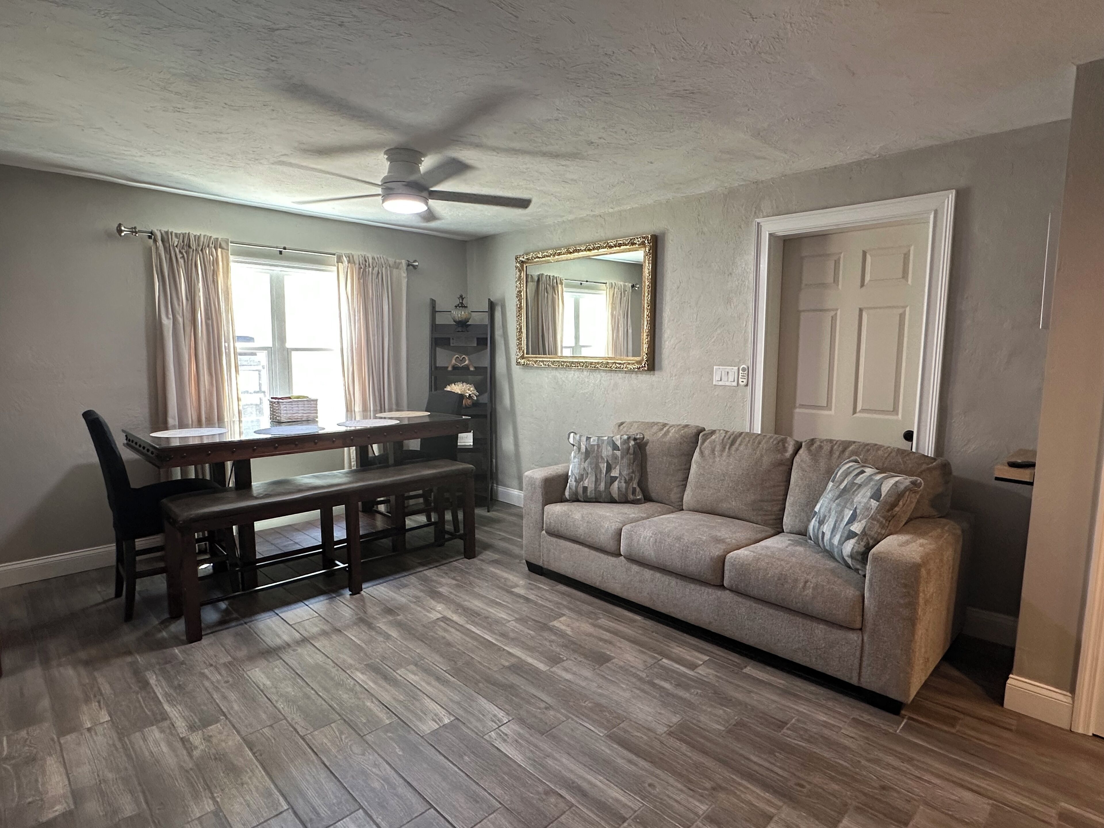 A dining area with a wooden table and chairs, a ceiling fan, and a gray sofa with decorative pillows.