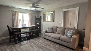 A dining area with a wooden table and chairs, a ceiling fan, and a gray sofa with decorative pillows.