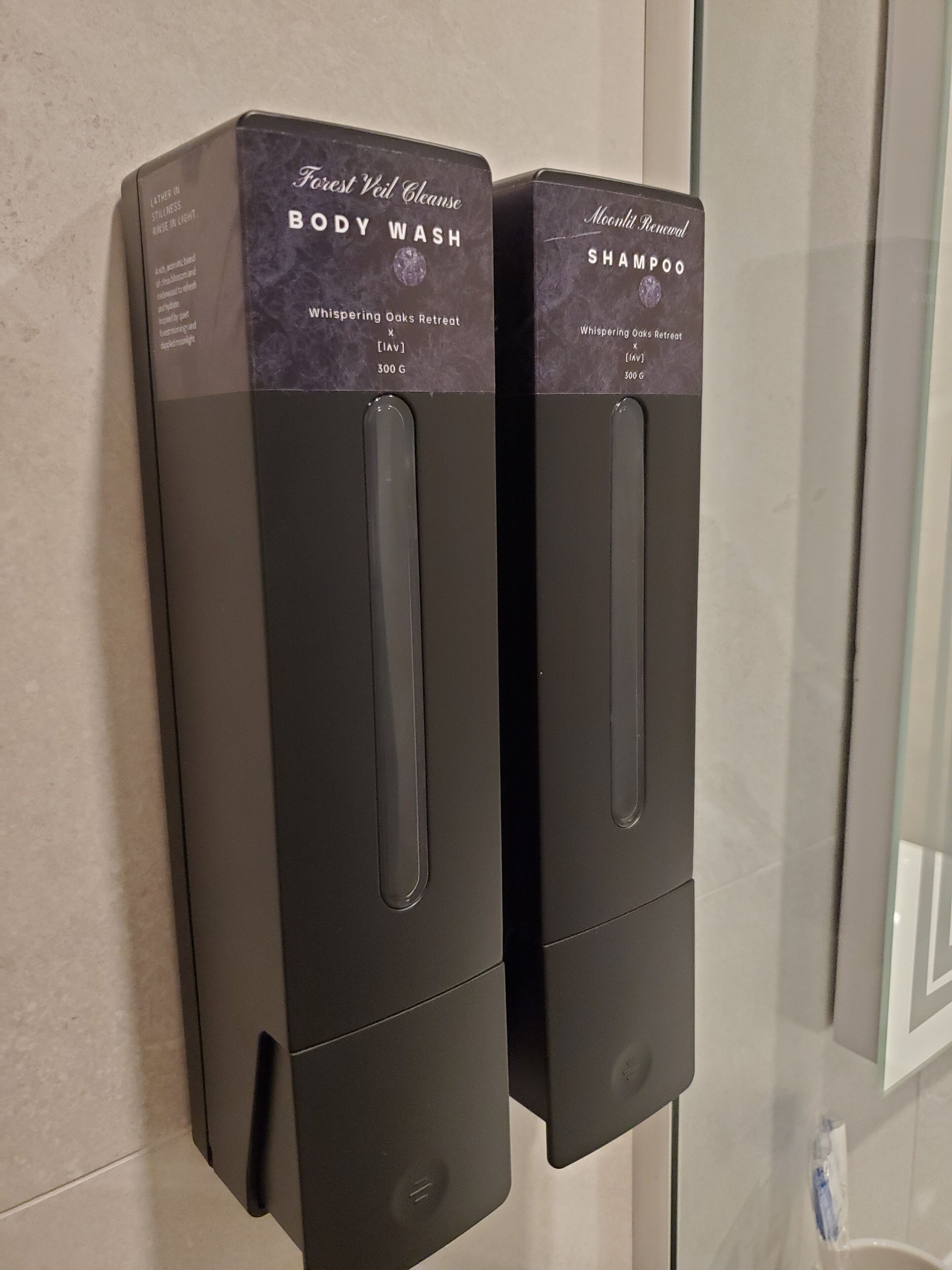 Two black dispensers labeled "Forest Veil Cleanse Body Wash" and "Moonlight Renewal Shampoo" in a bathroom.