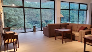 A spacious living room with large windows offering a view of a forested area. There is a sofa, a small table, and a chair.