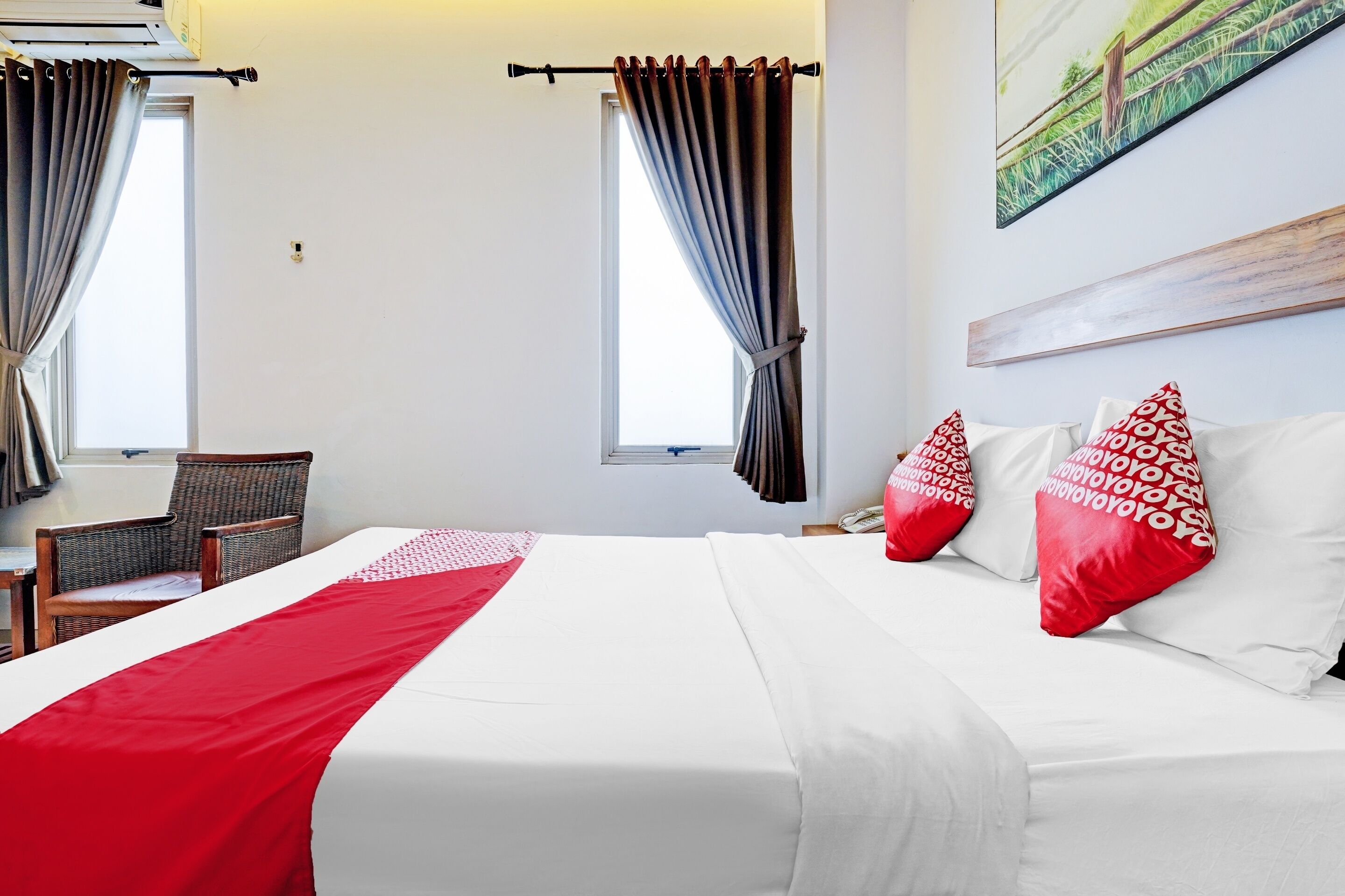 A hotel room with a bed, two windows with curtains, a chair, and a painting on the wall.